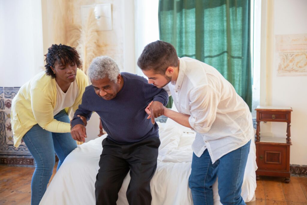 Two people helping an elderly man stand up from a bed.